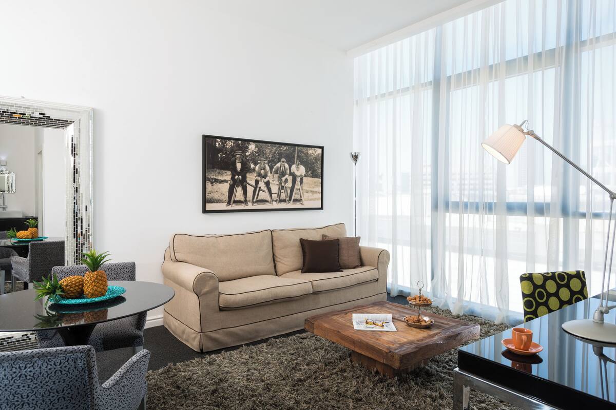 Un salon moderne avec un canapé beige, une table basse et un coin repas comprenant des chaises et une table. On y trouve une grande fenêtre avec des rideaux légers, un tableau encadré au mur et un lampadaire.