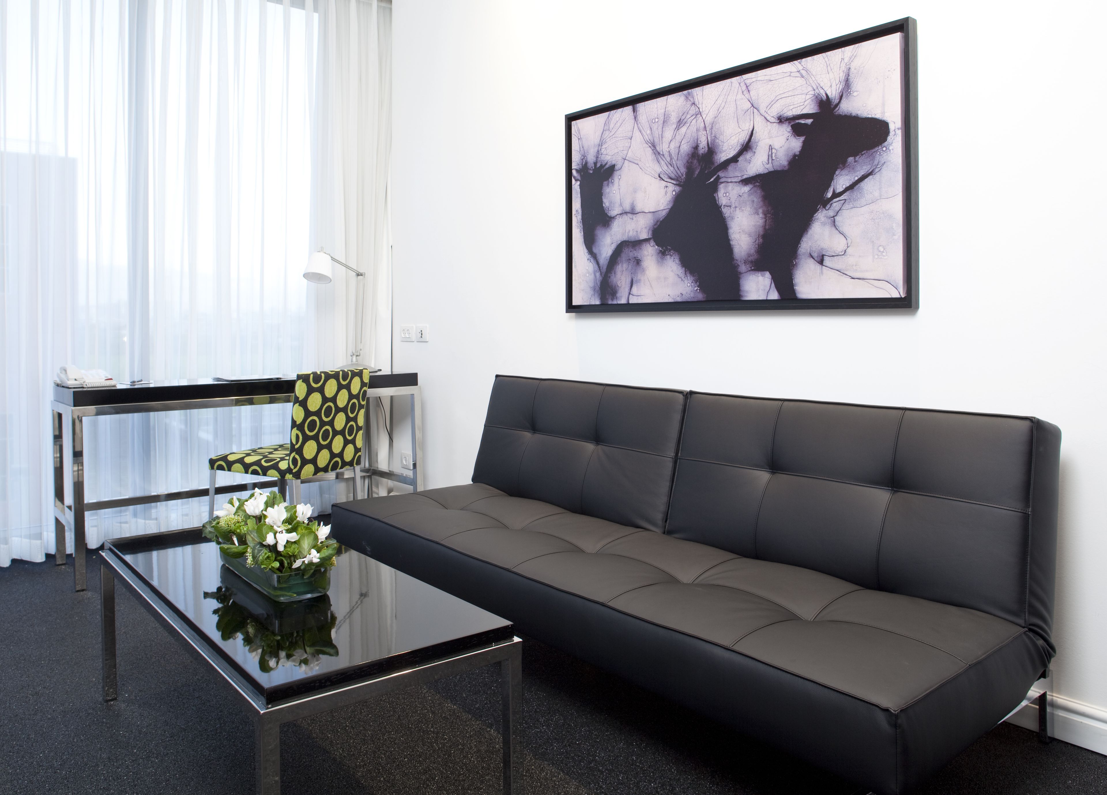 A modern living room with a glass coffee table, a patterned armchair, a grey sofa, and a large abstract painting on the wall.