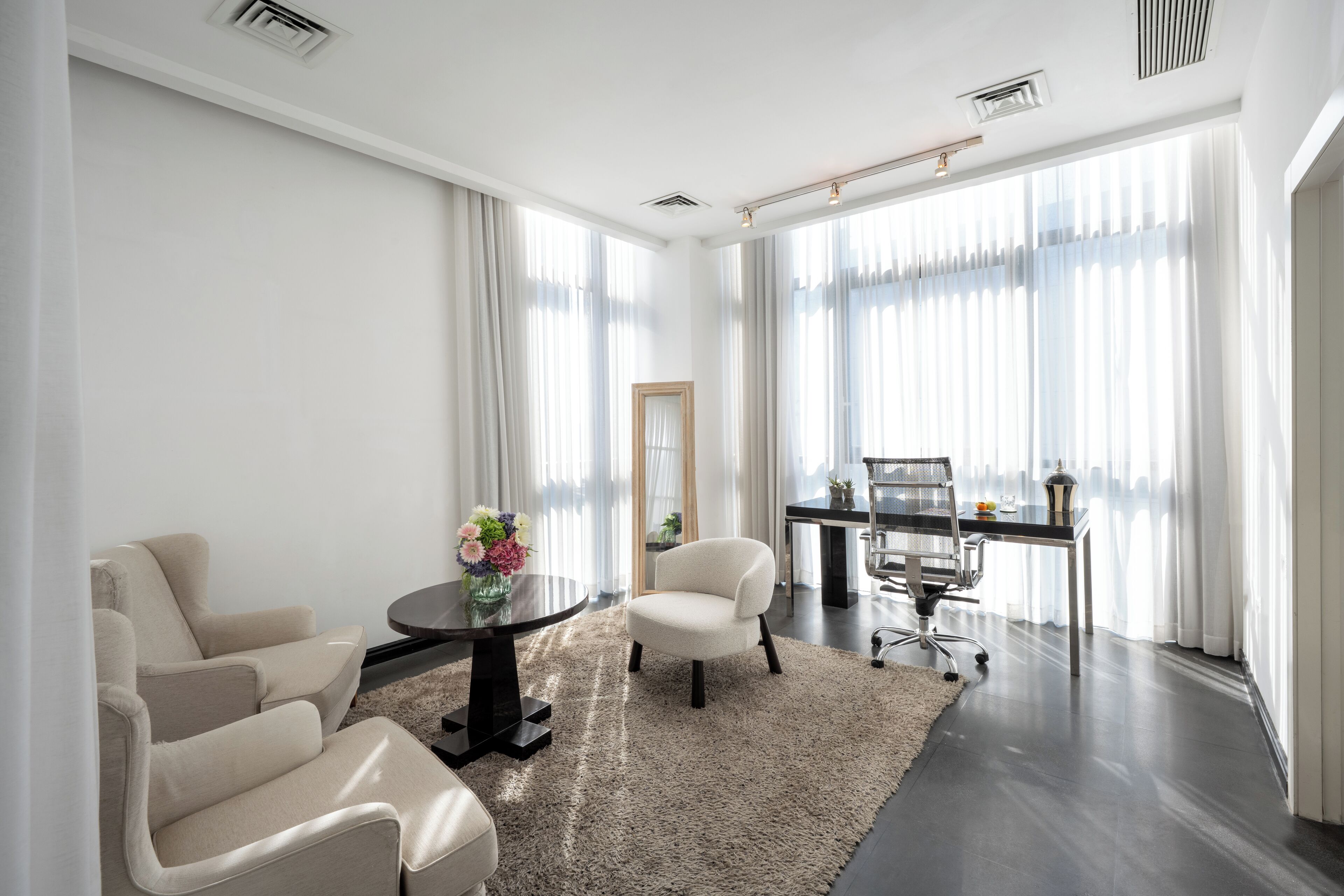 Une salle de réunion moderne, dotée d’une table ronde, de deux chaises, d’un bureau avec un ordinateur et de grandes fenêtres ornées de rideaux légers.
