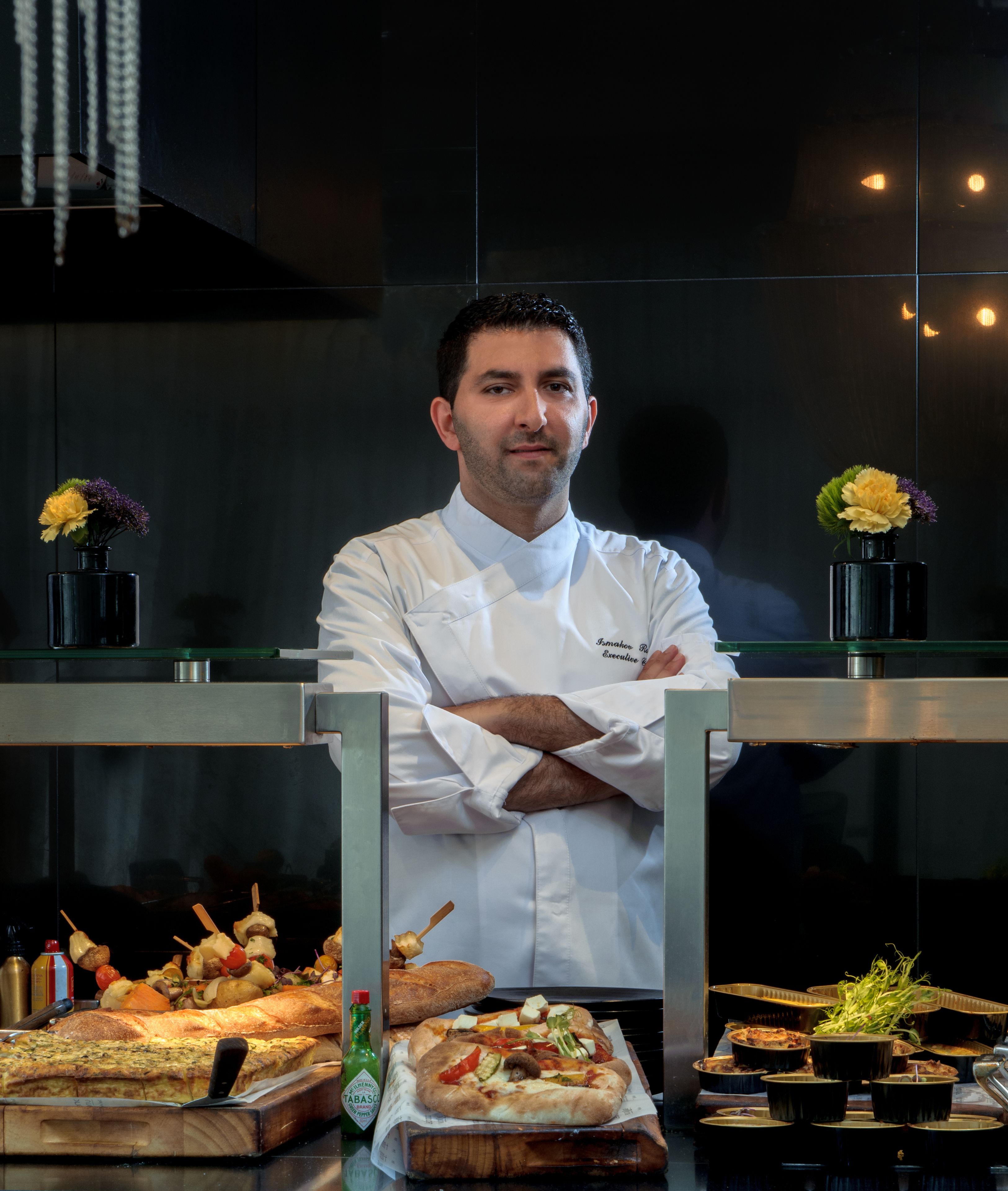 A chef standing in a kitchen with a variety of dishes and a bottle of Tabasco sauce.