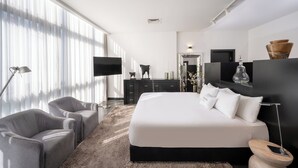 Un dormitorio moderno con una cama grande, dos sillones grises, un aparador oscuro y un televisor de pantalla plana.