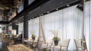 Un vestíbulo amplio con diseño moderno, que incluye un gran área de estar con sillas blancas y plantas en macetas, una alfombra estampada y un techo alto con vigas a la vista.
