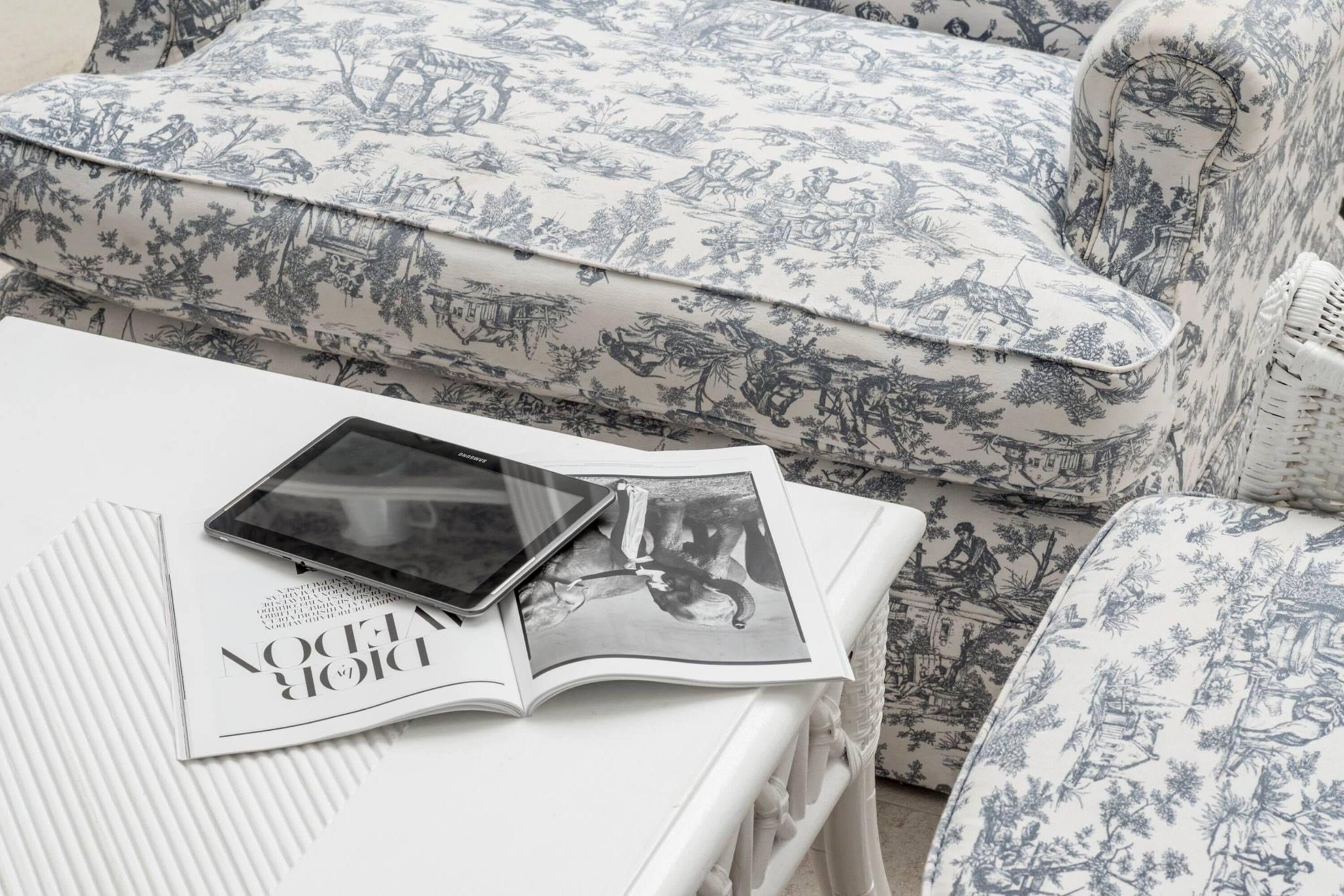 A white sofa with a blue and white floral pattern, a tablet on a magazine, and a wicker chair with a similar pattern.