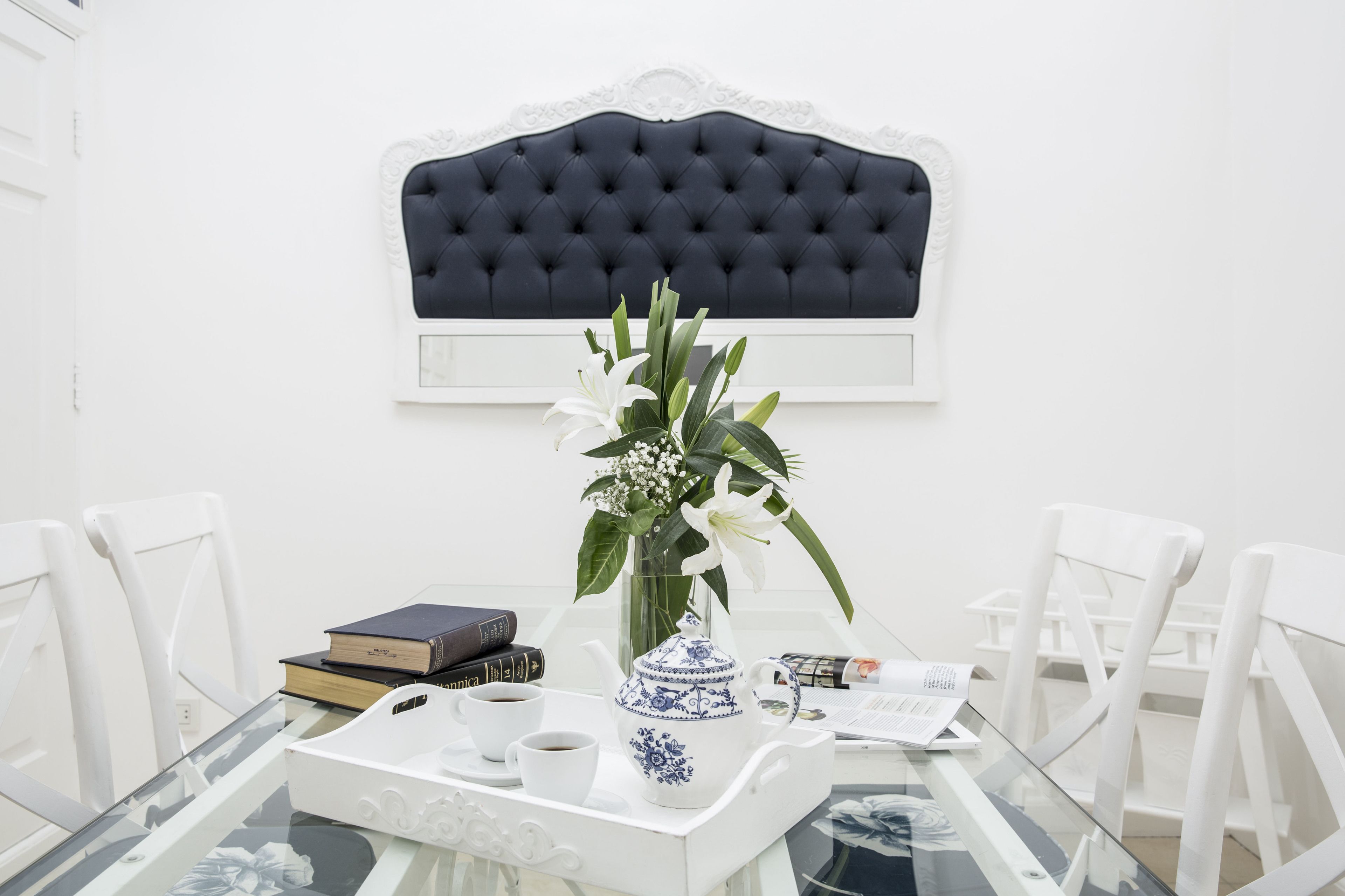 A dining table with a glass top, white chairs, a vase with flowers, a teapot, and books.