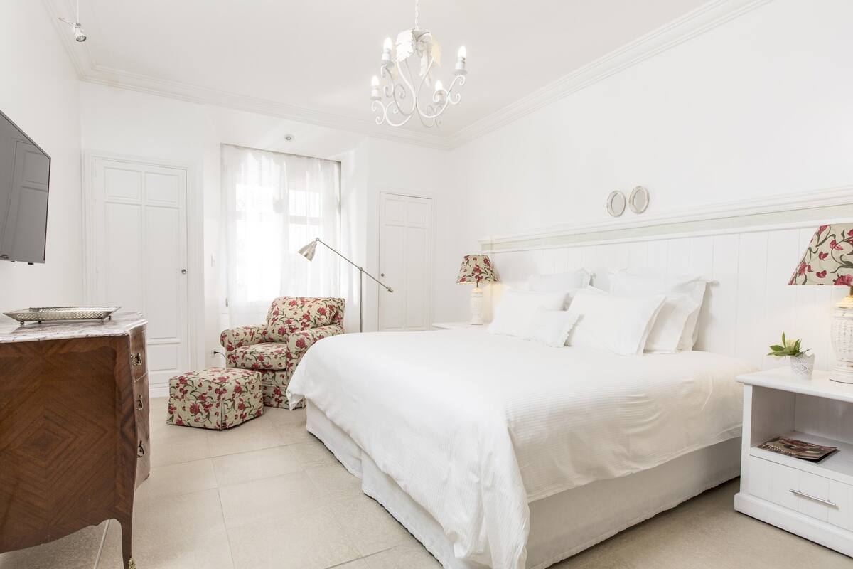 Un dormitorio con decoración en tonos blancos, una cama grande, una mesita de noche de madera, un sillón con estampado floral y una araña.
