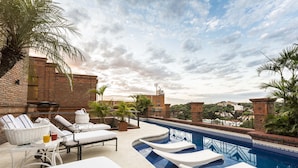 A rooftop pool area with lounge chairs, a table with a basket and drinks, and a view of the cityscape.
