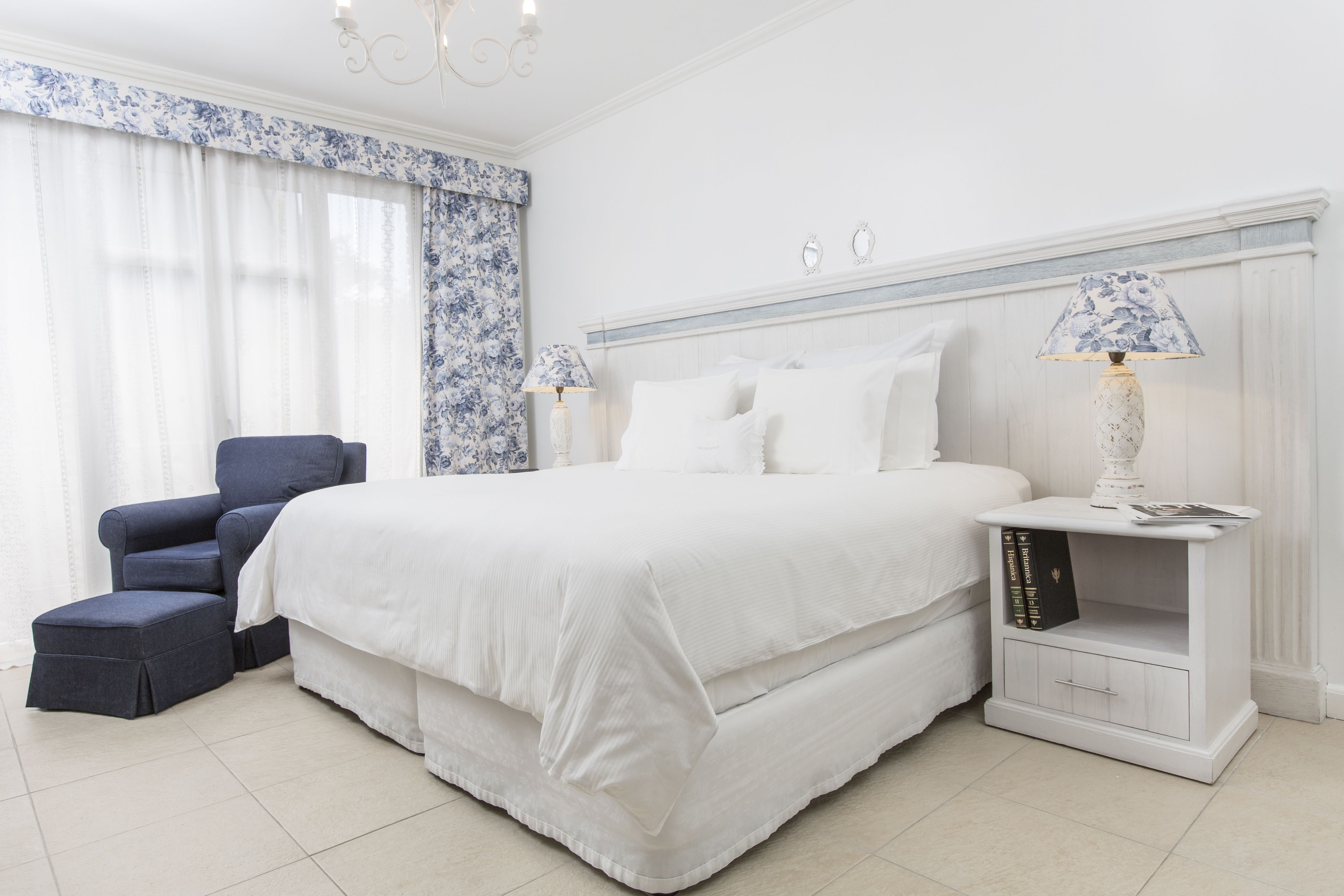 A bedroom with a large bed, a chair, a nightstand with a lamp, and a window with curtains.
