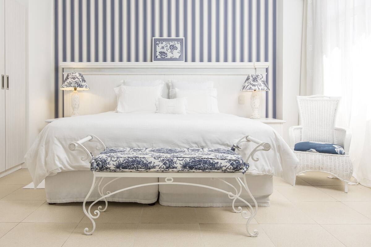 Un dormitorio ordenado con una cama grande, lámparas de noche, un cojín decorativo, un banco y una silla de mimbre.