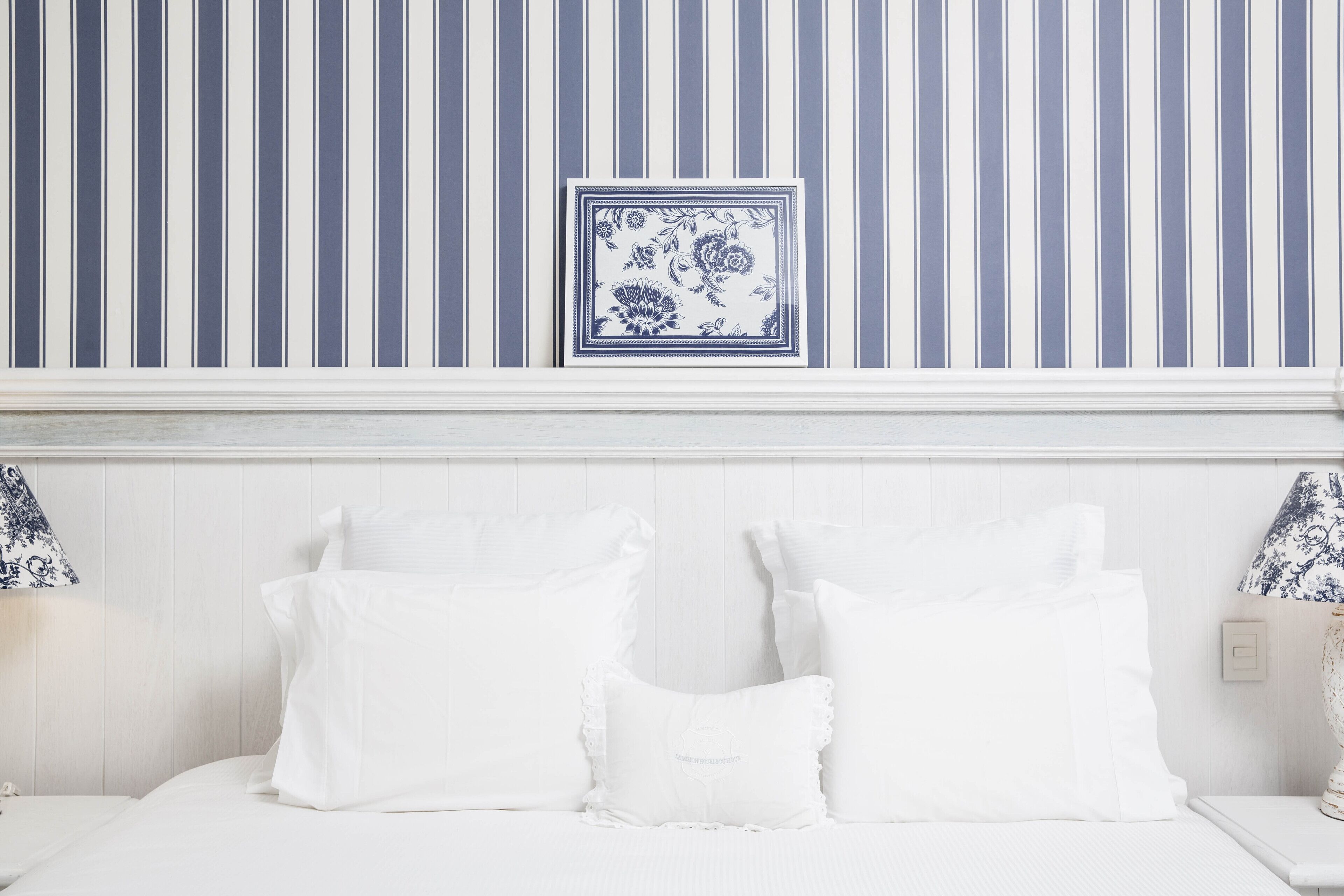 Una cama blanca con almohadas blancas, una pared azul y blanca a rayas y una obra de arte floral azul y blanca enmarcada sobre la cama.