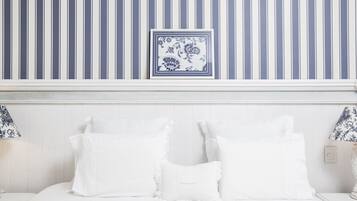 Una cama blanca con almohadas blancas, una pared azul y blanca a rayas y una obra de arte floral azul y blanca enmarcada sobre la cama.