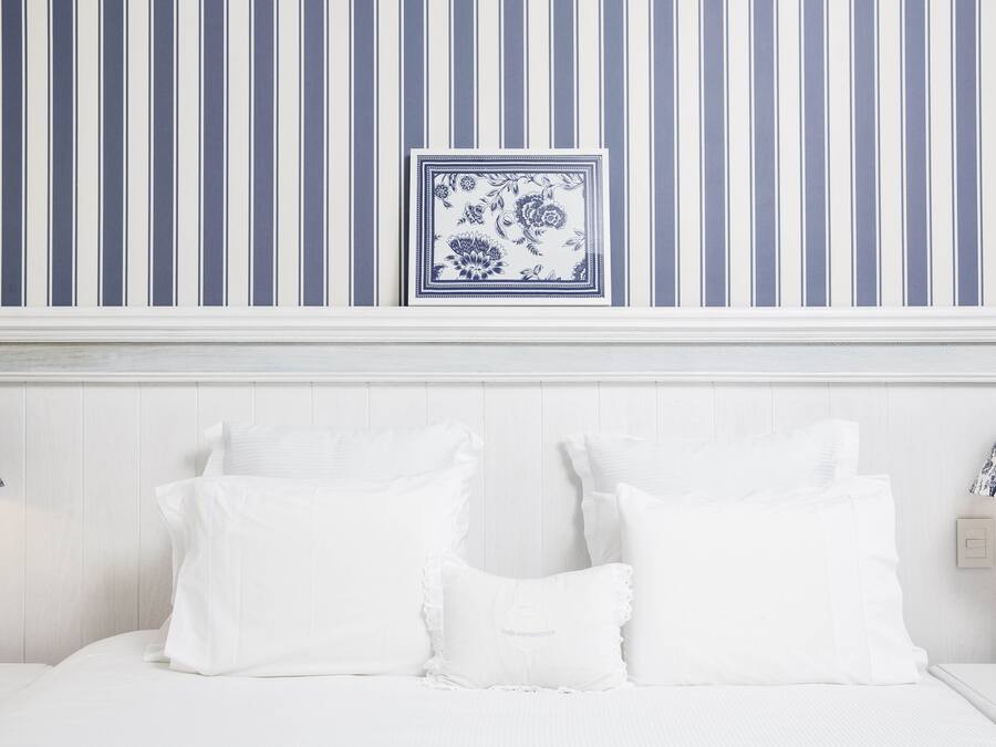 Una cama blanca con almohadas blancas, una pared azul y blanca a rayas y una obra de arte floral azul y blanca enmarcada sobre la cama.