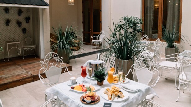An outdoor dining area with white metal chairs and tables, surrounded by potted plants and a wooden deck.