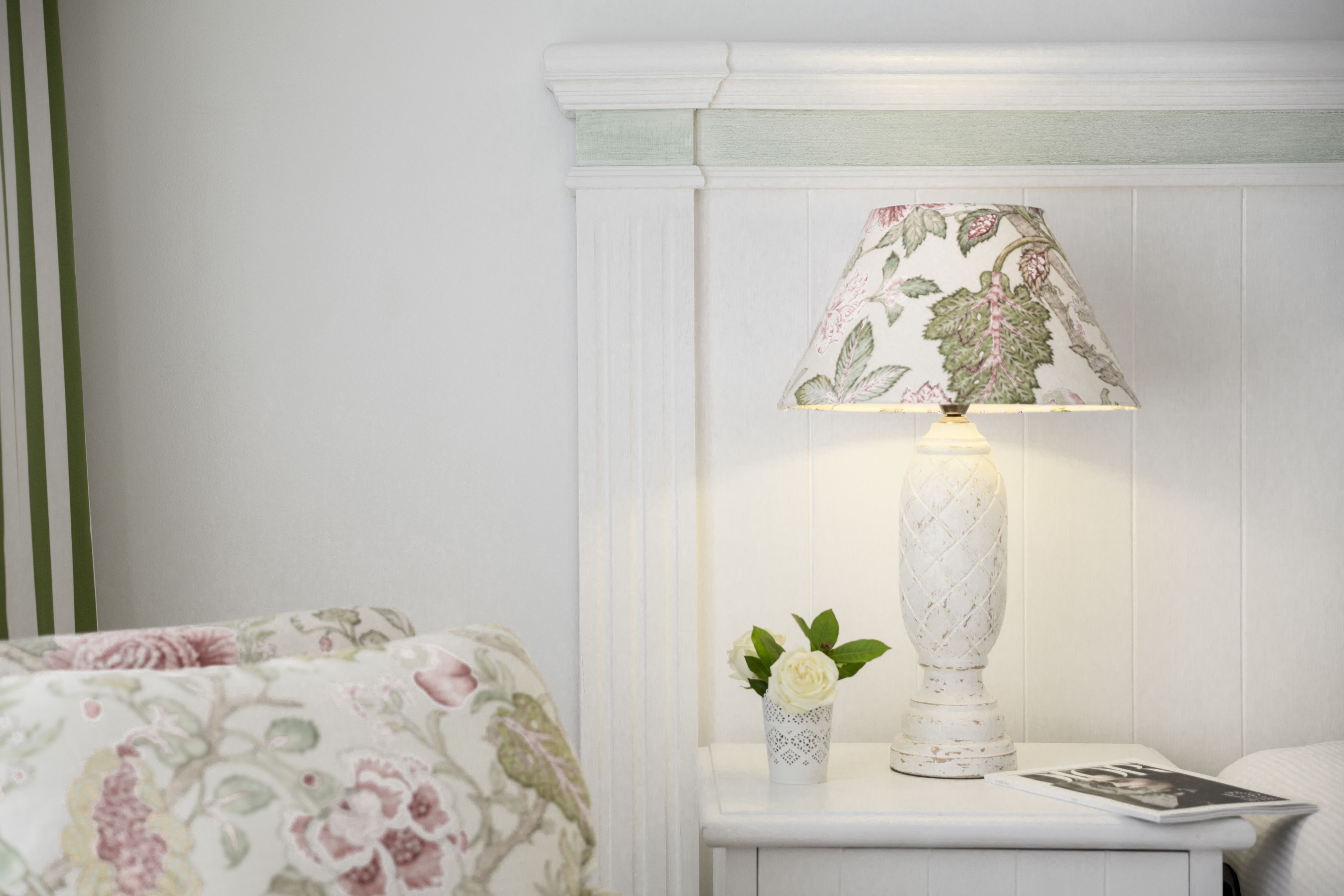 Uma cama bem arrumada com roupa de cama florida, uma mesa de cabeceira com abajur e um pequeno vaso com uma única flor, e uma revista sobre a mesa.