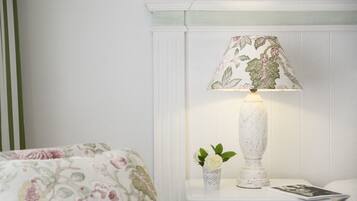 Uma cama bem arrumada com roupa de cama florida, uma mesa de cabeceira com abajur e um pequeno vaso com uma única flor, e uma revista sobre a mesa.