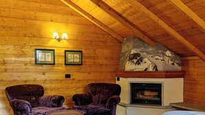 Ein Blockhauszimmer mit zwei violetten Sesseln, einem kleinen runden Tisch, einem Kamin mit Steinverkleidung und gerahmten Pflanzenabbildungen an der Wand.