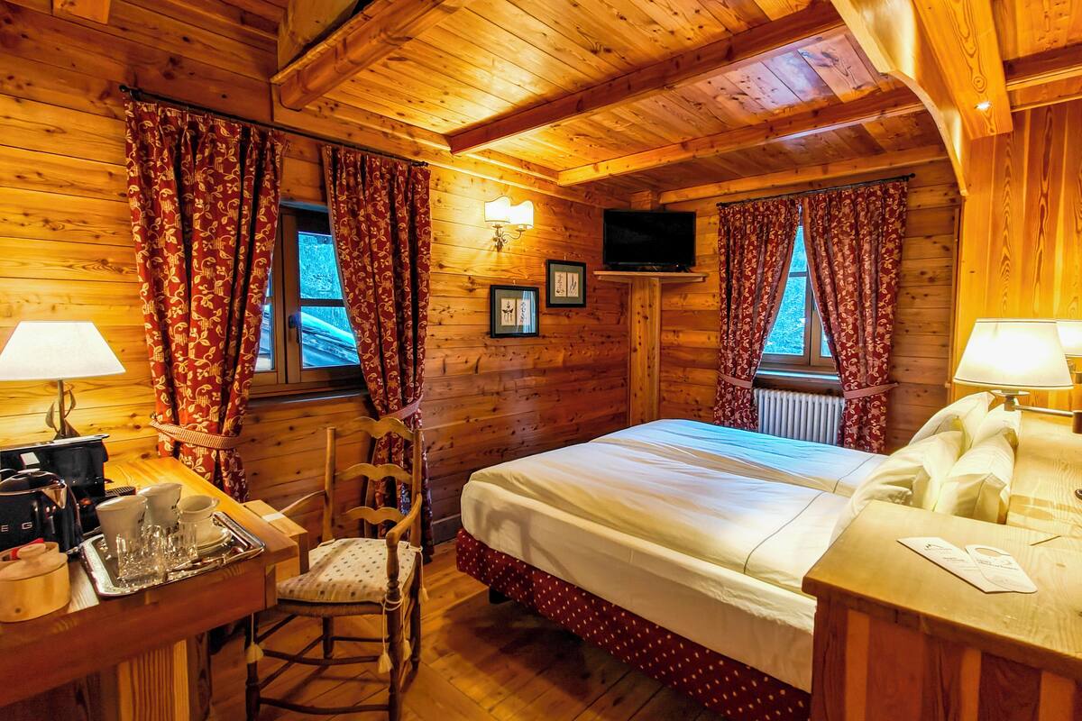 A cozy cabin room with wooden walls, a bed, a chair, a desk with a lamp, and a TV.