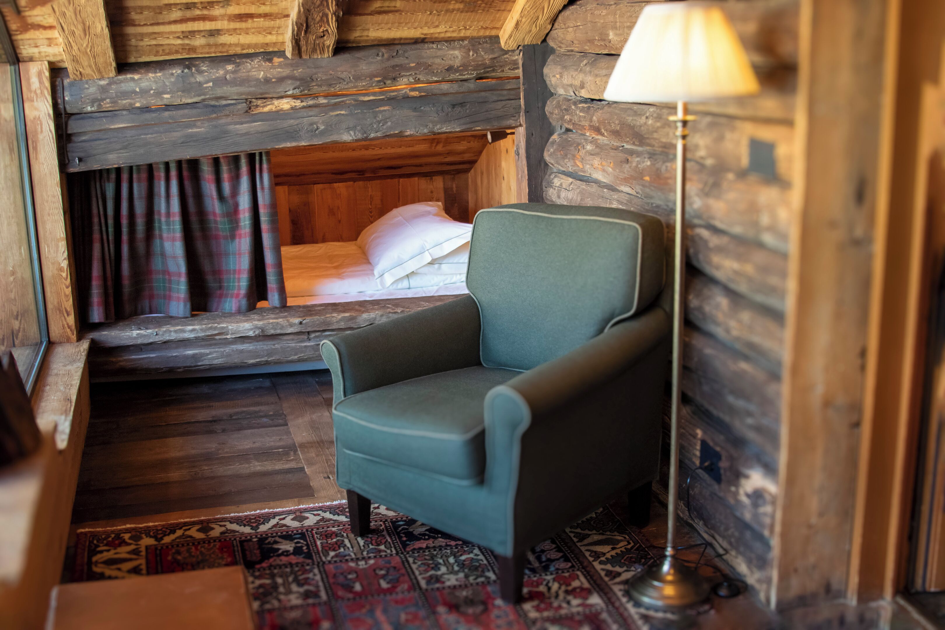 Une chambre chaleureuse dans un chalet en rondins, avec un lit simple, un fauteuil vert, un tapis à motifs et une lampe.
