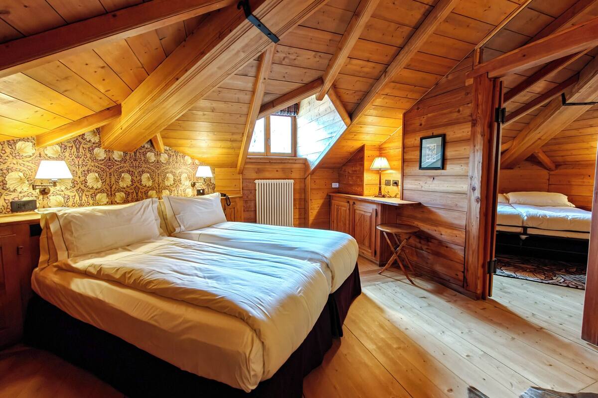 A cozy attic bedroom with a wooden ceiling, a bed with white linens, a desk with a lamp, and a radiator.