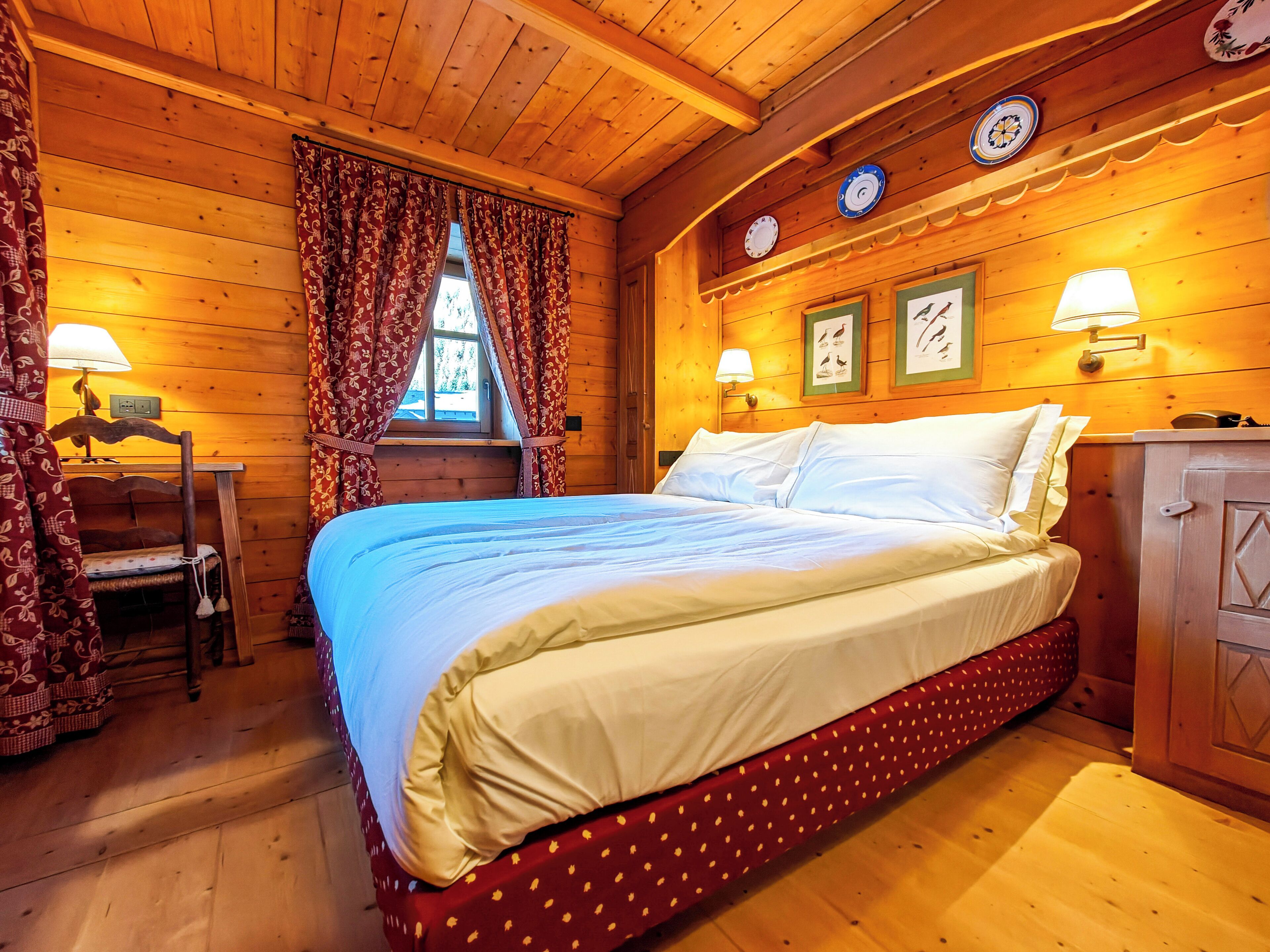 Ein Schlafzimmer einer Holzhütte mit Bett, Vorhängen, einem Fenster, Wanduhren und einer Lampe.