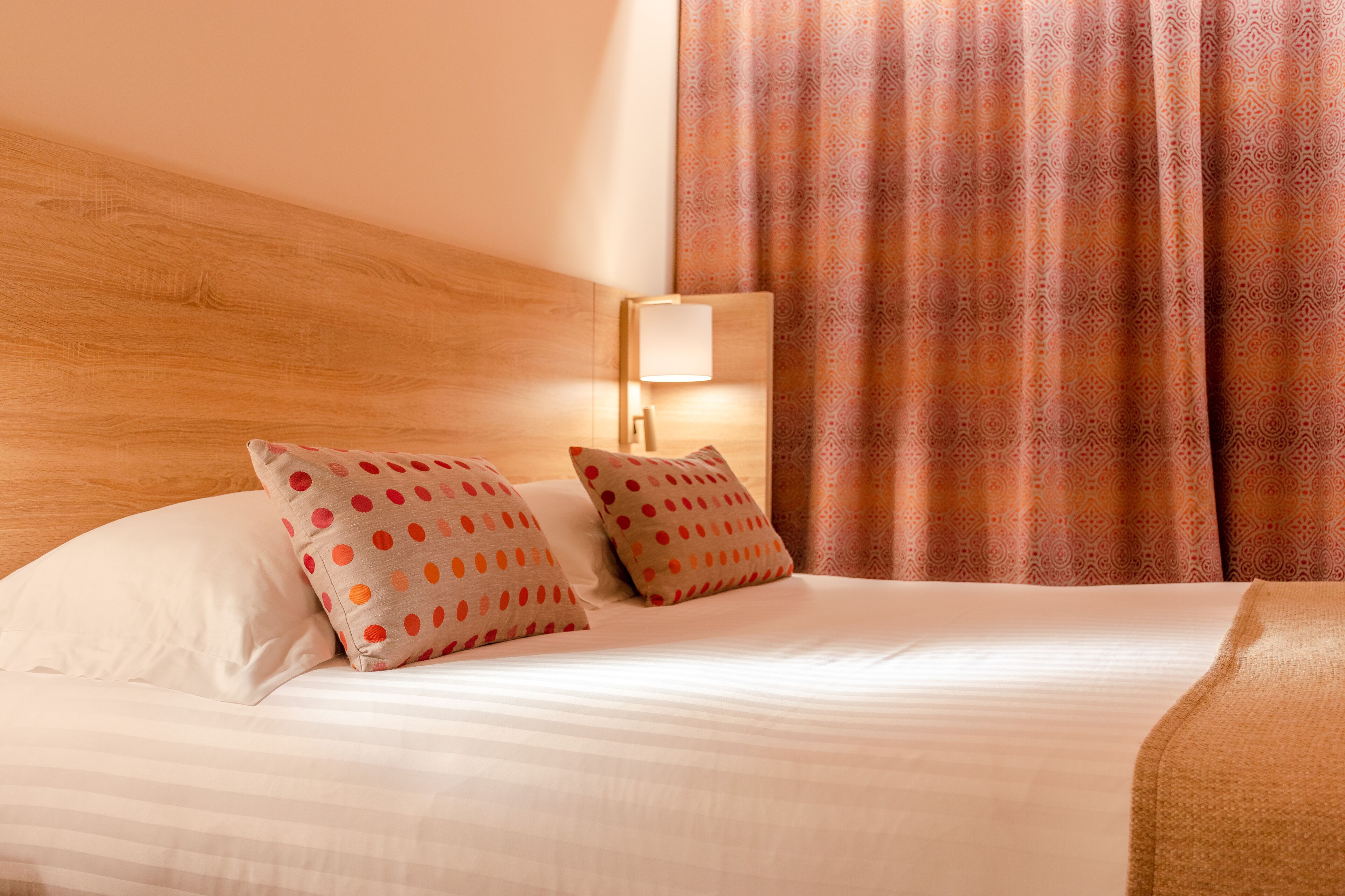 Una cama bien hecha con dos almohadas, una lámpara de noche y una cortina estampada.