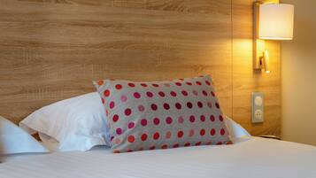 Una cama bien tendida con sábanas blancas y un cojín de lunares, un cabecero de madera y una lámpara fijada a la pared.