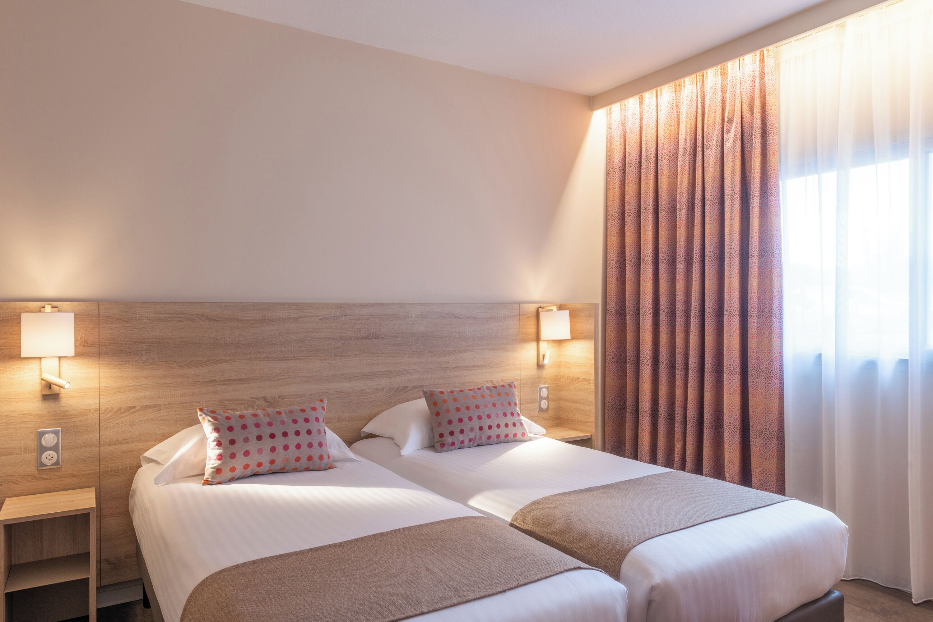 A double bed with two pillows, a nightstand with a lamp, and a window with patterned curtains.