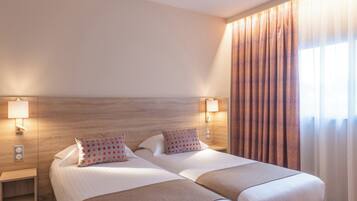 A double bed with two pillows, a nightstand with a lamp, and a window with patterned curtains.