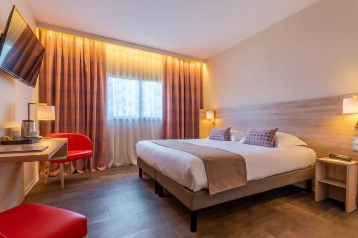Una habitación de hotel con una cama, un escritorio, una silla, un televisor y un ventanal con cortinas.