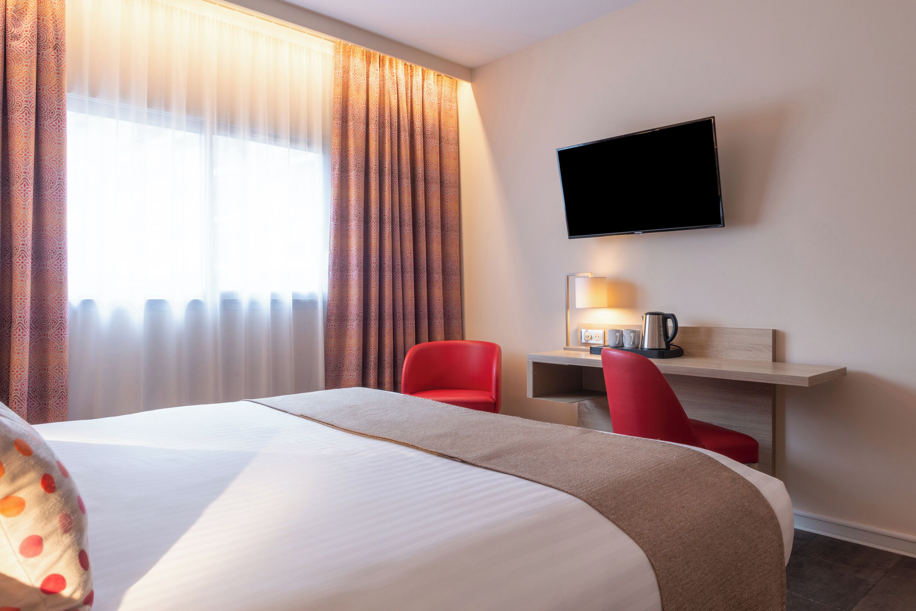 A hotel room with a large bed, a desk with a lamp, a red chair, and a television on the wall.