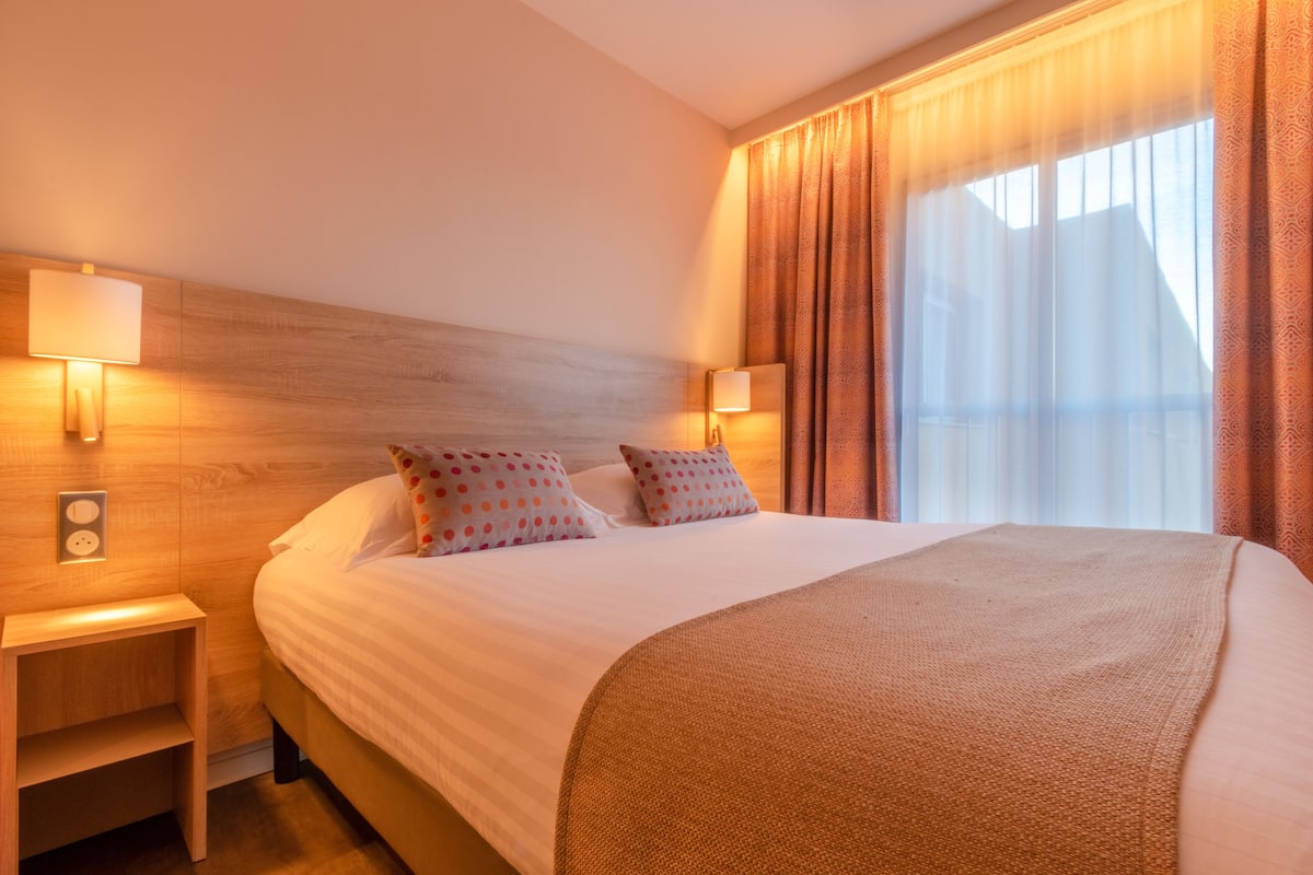 Una habitación de hotel con una cama grande, dos lámparas de noche y un ventanal con cortinas.