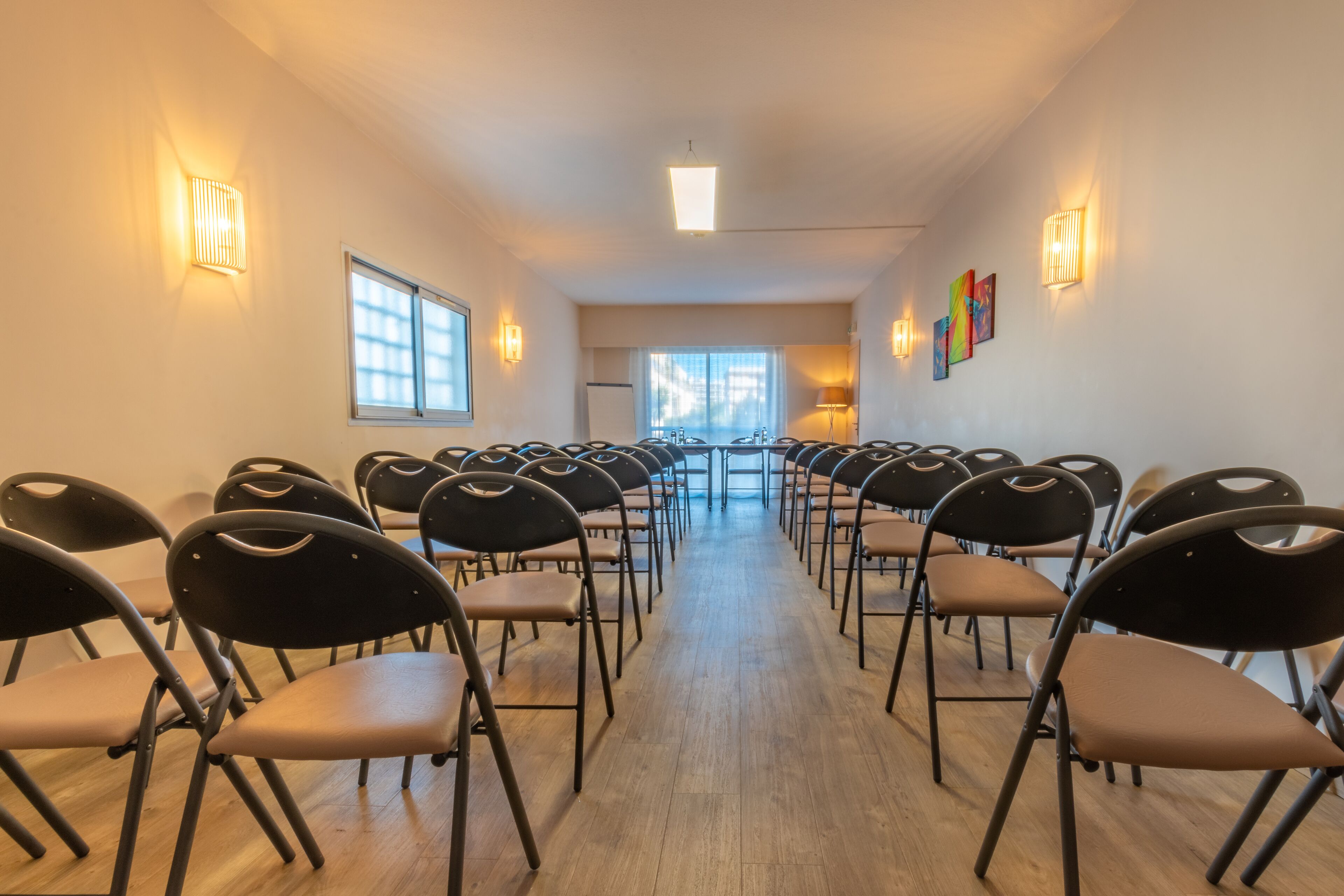 Une salle de conférence avec des rangées de chaises disposées face à une scène, équipée de tables et de chaises, et éclairée par des appliques murales.