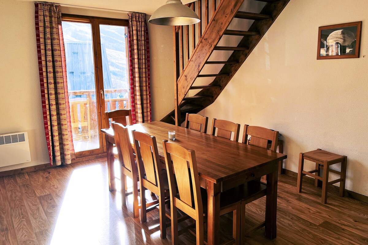 Un comedor con una mesa y sillas de madera, una escalera y una ventana con cortinas.