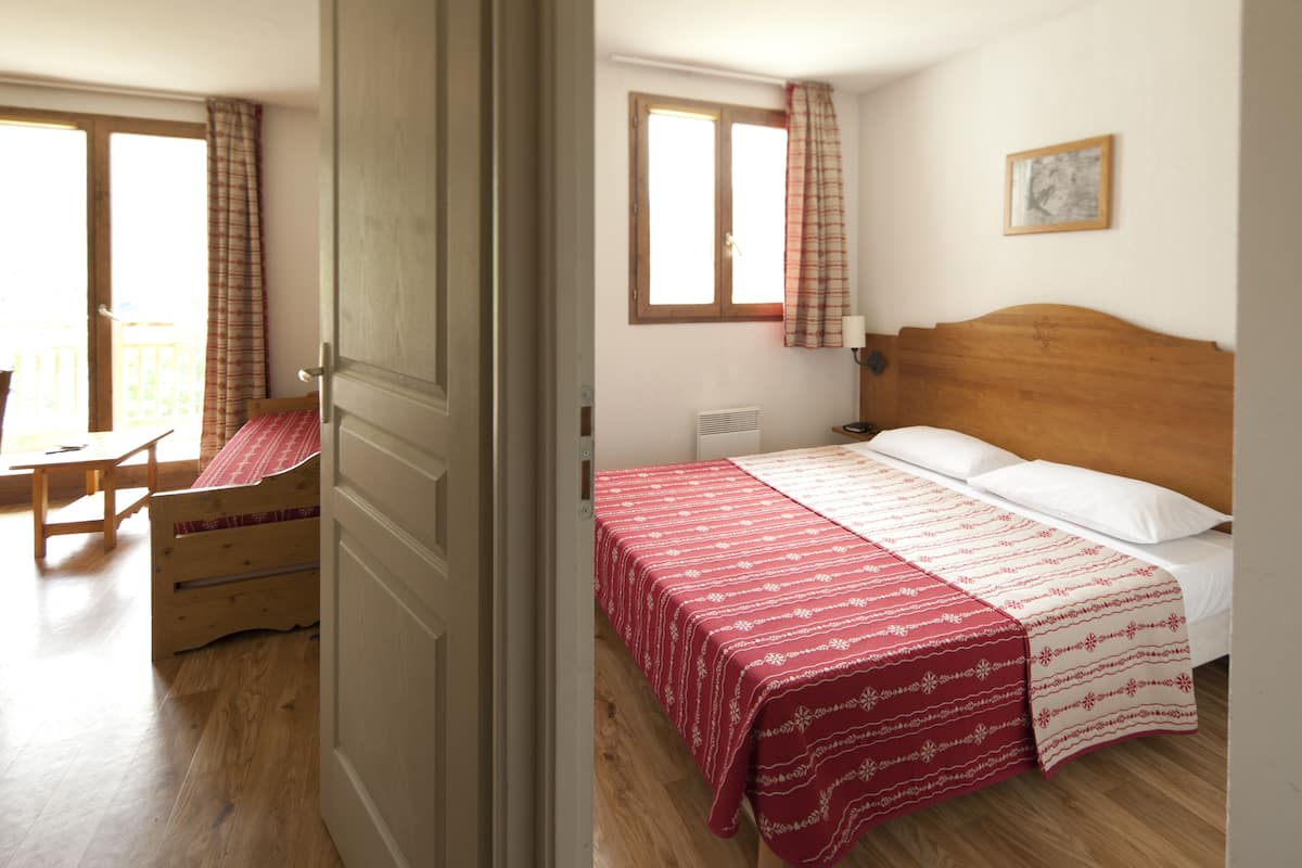 Un dormitorio con una cama de madera, una colcha roja y blanca, una mesita de noche de madera, una mesita auxiliar y una ventana con cortinas.