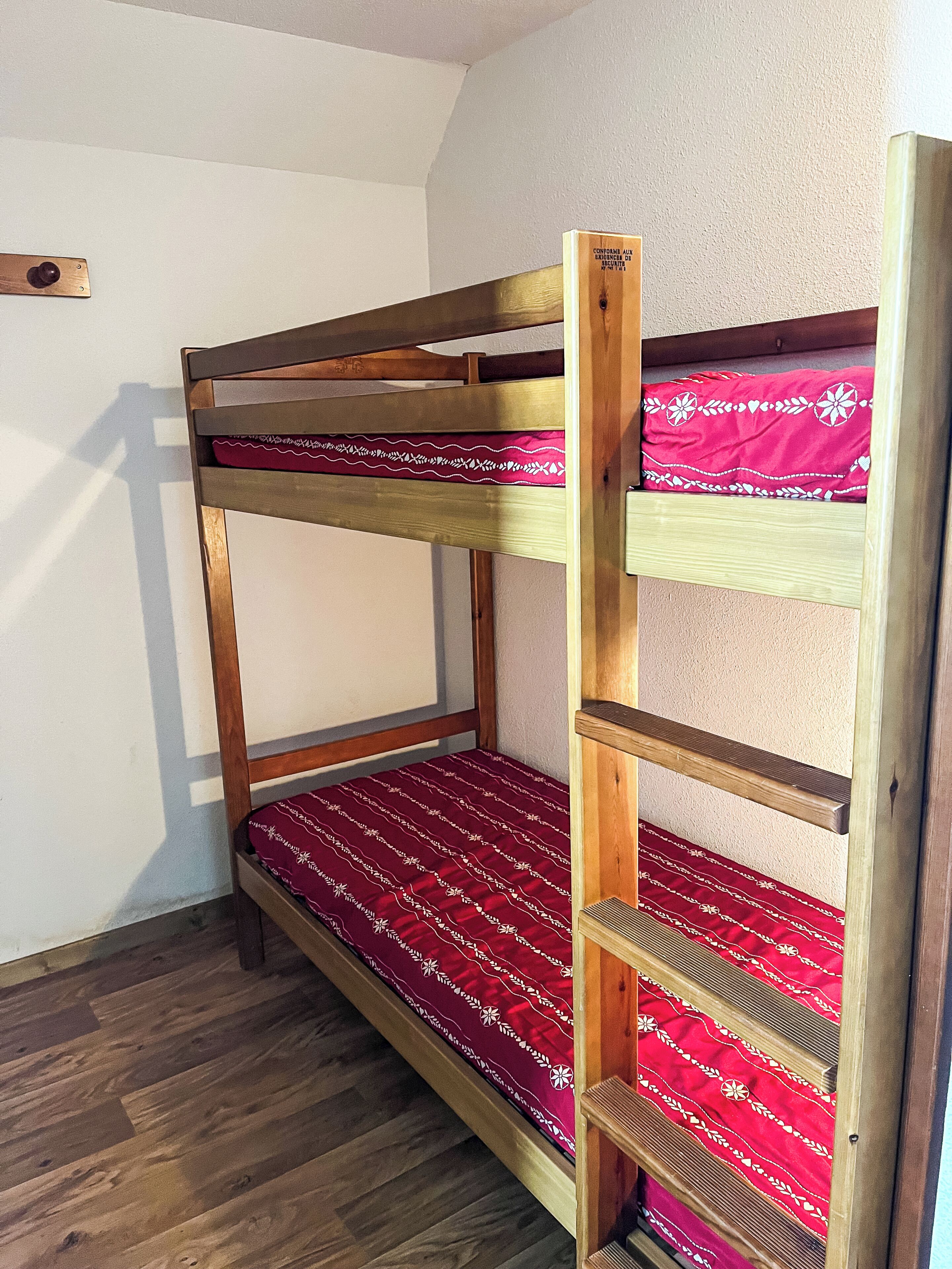A bunk bed with a red patterned bedspread, wooden frame, and a shelf on the lower bunk.
