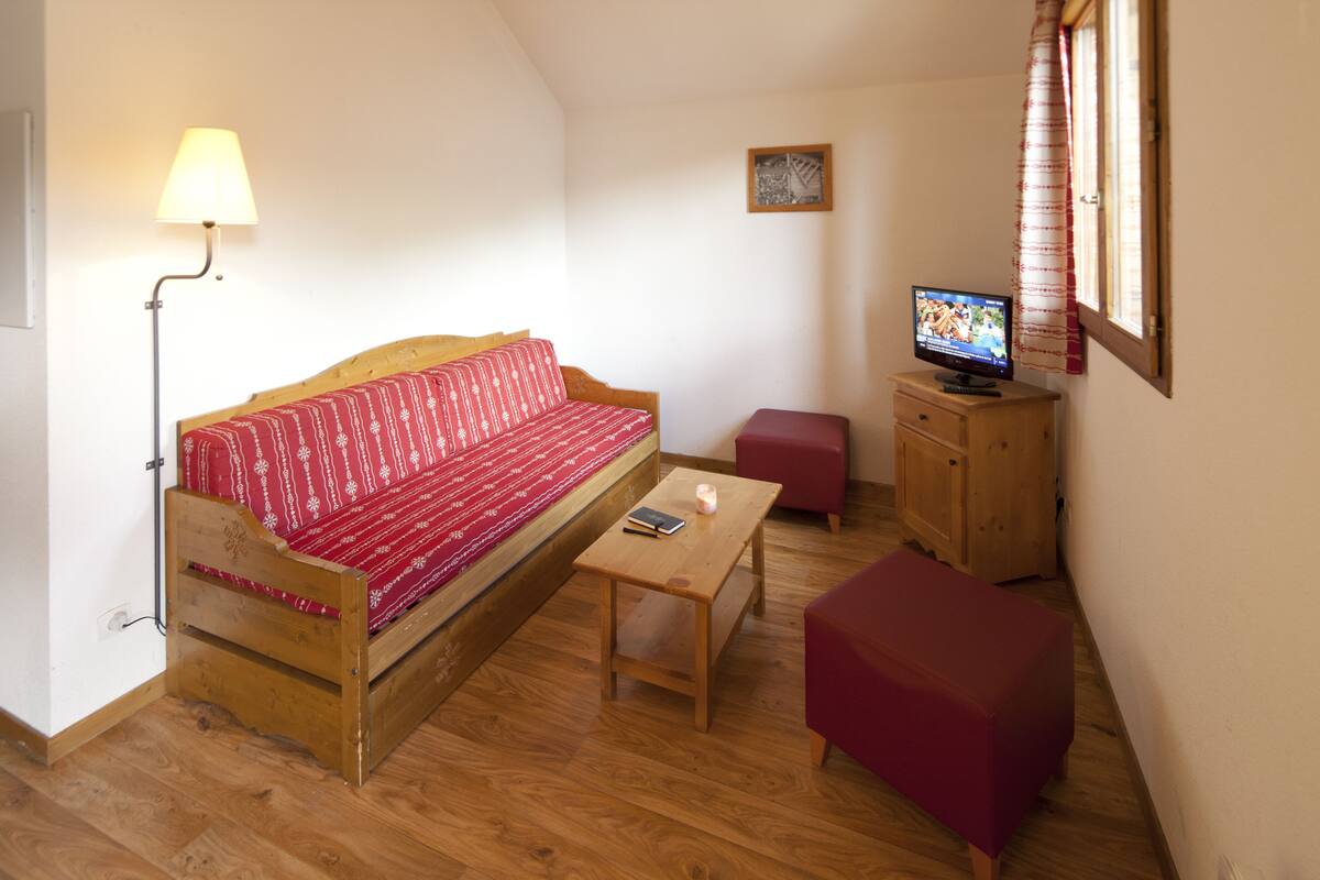 Una habitación acogedora con un sofá de madera, una mesita pequeña de madera, un reposapiés rojo, un televisor sobre un mueble de madera y una lámpara de pie.