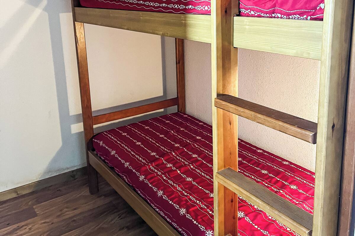 Una cama litera con un edredón rojo estampado, estructura de madera y un estante en la parte inferior.