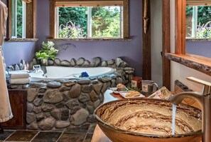 A bathroom with a stone wall, a bathtub, a wooden basin, and a window with a view of greenery.