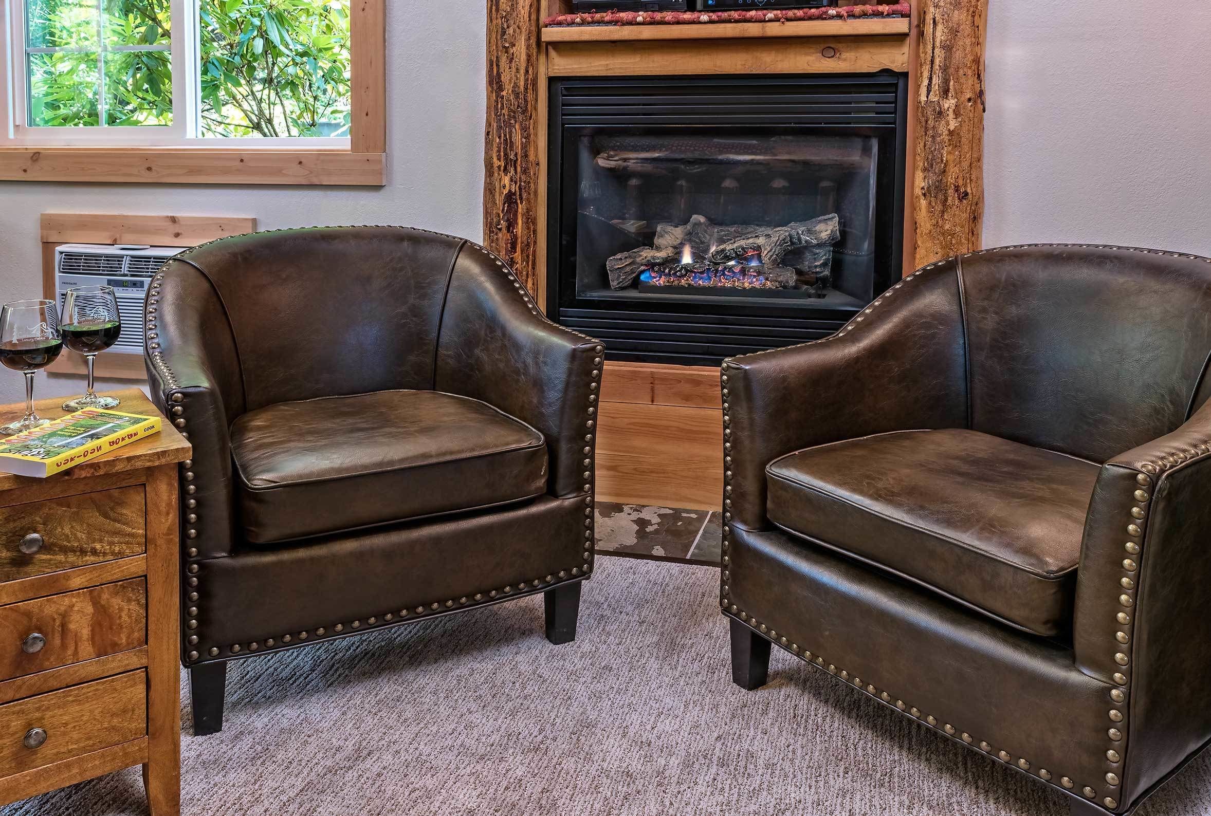Deux fauteuils en cuir ornés de clous en métal, une table d’appoint en bois avec un livre et des lunettes, et une cheminée avec des bûches.