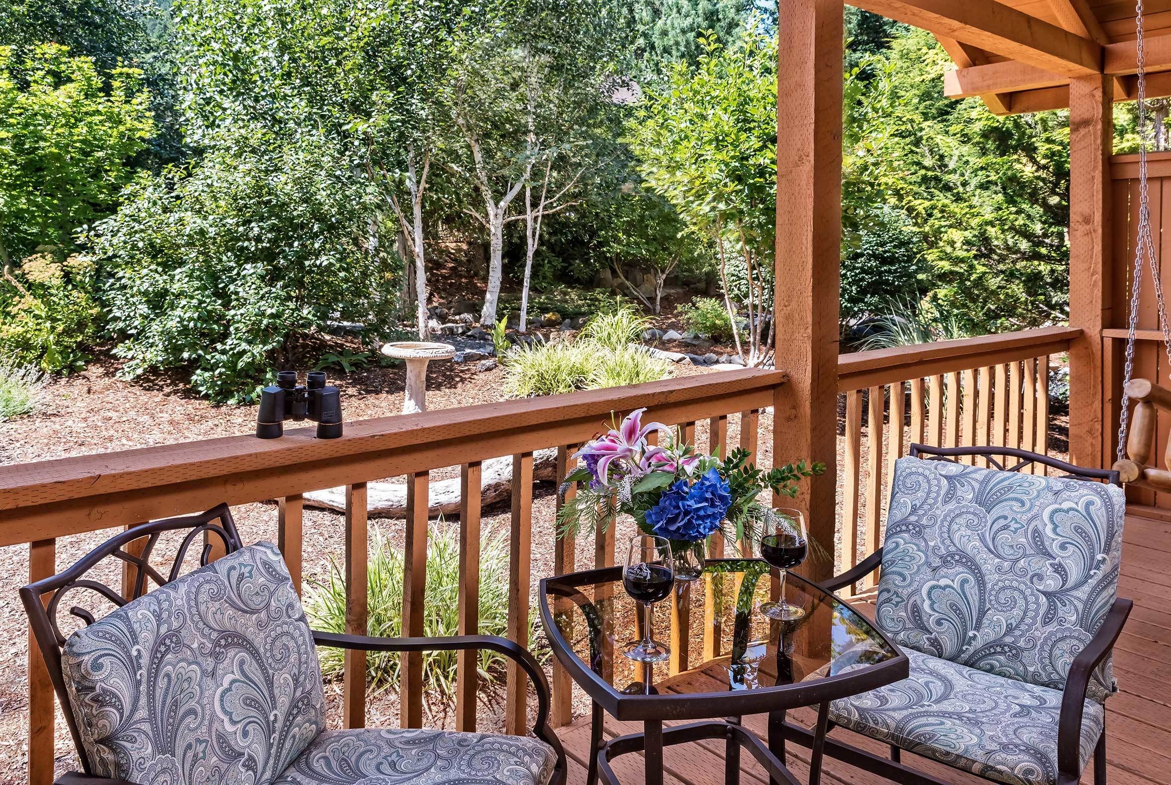 An outdoor deck with wooden railings, two patterned chairs, a small table with wine glasses and a vase of flowers.