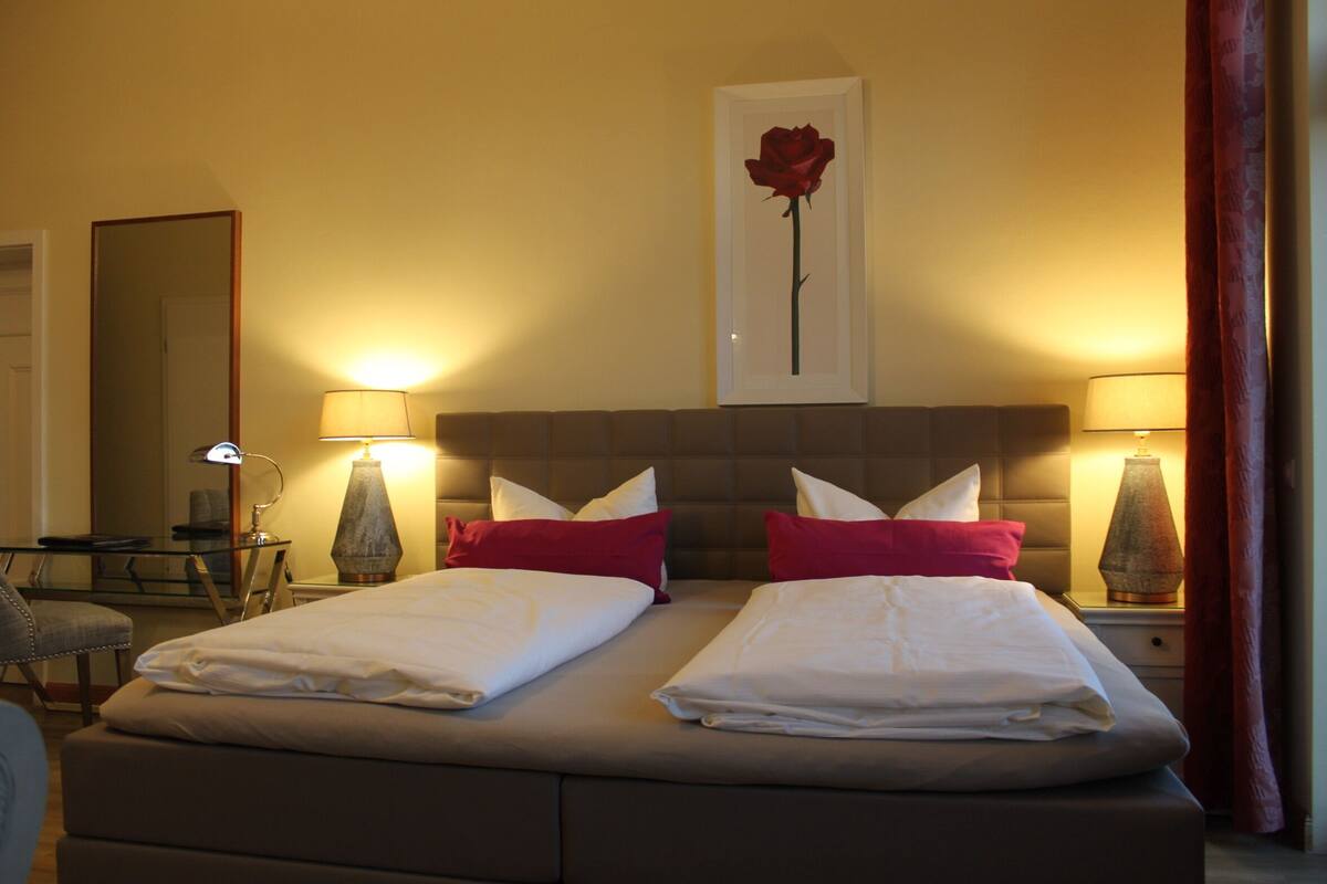 A double bed with white linens, two red pillows, and a framed rose on the wall.