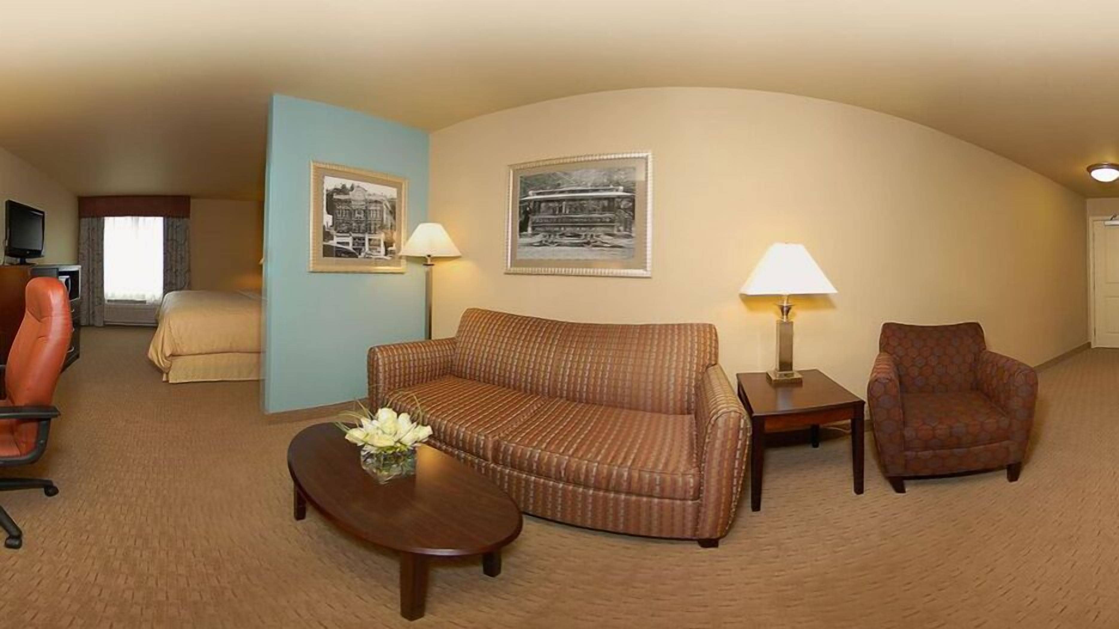 Habitación de hotel con sofá, sillón y una mesita de noche con lámpara. Se observa un televisor y una ventana con cortinas.