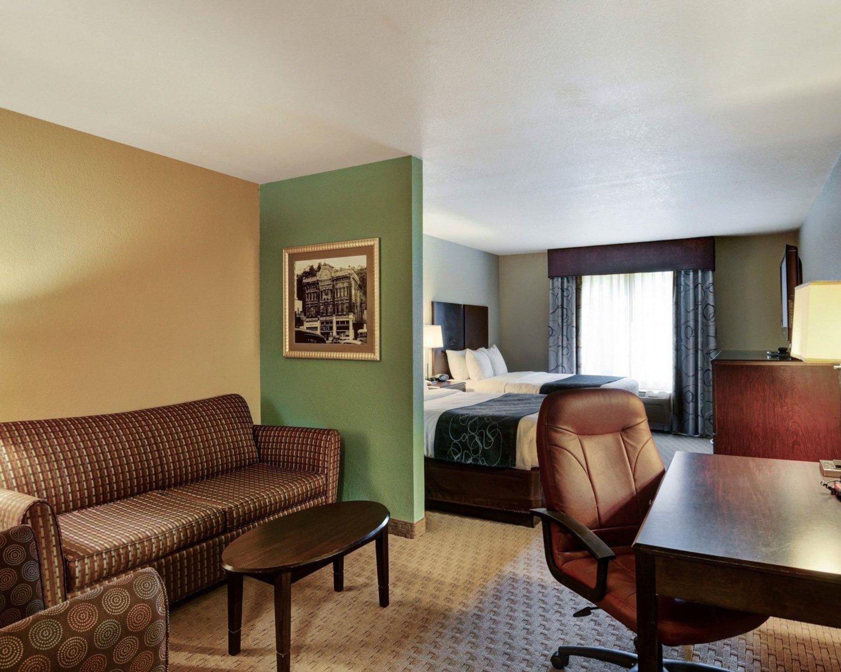 Habitación de hotel con sofá, sillón y una pequeña mesa. Se ve una cama con cabecera y una ventana con cortinas. En la pared hay un cuadro enmarcado.