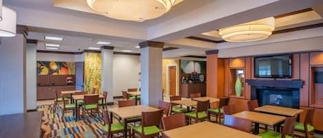 A hotel dining area with multiple tables and chairs, a television mounted on the wall, and a patterned carpet.