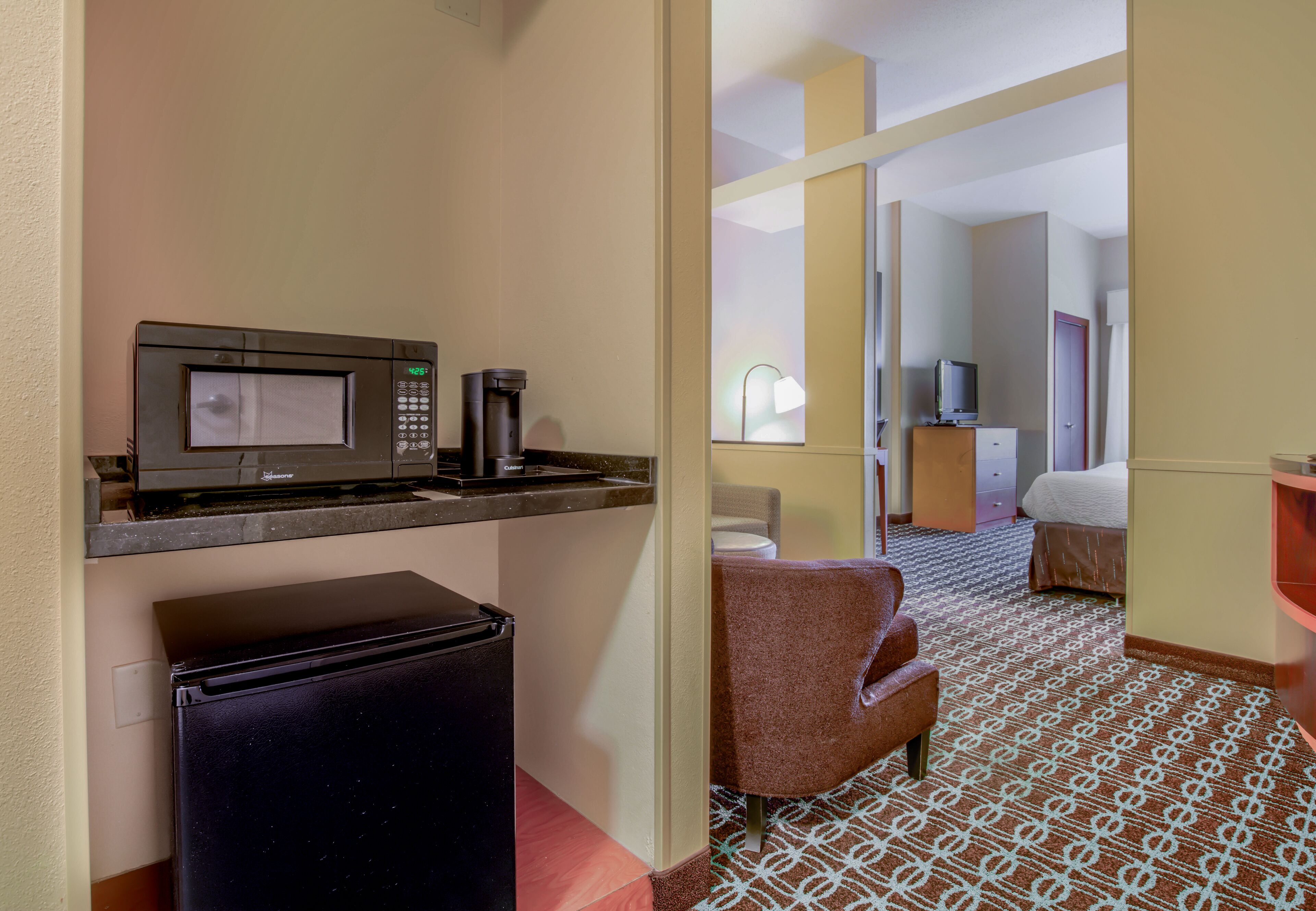 A hotel room with a kitchenette including a microwave, coffee maker, and refrigerator. There is a brown armchair and a bed with a patterned carpet.