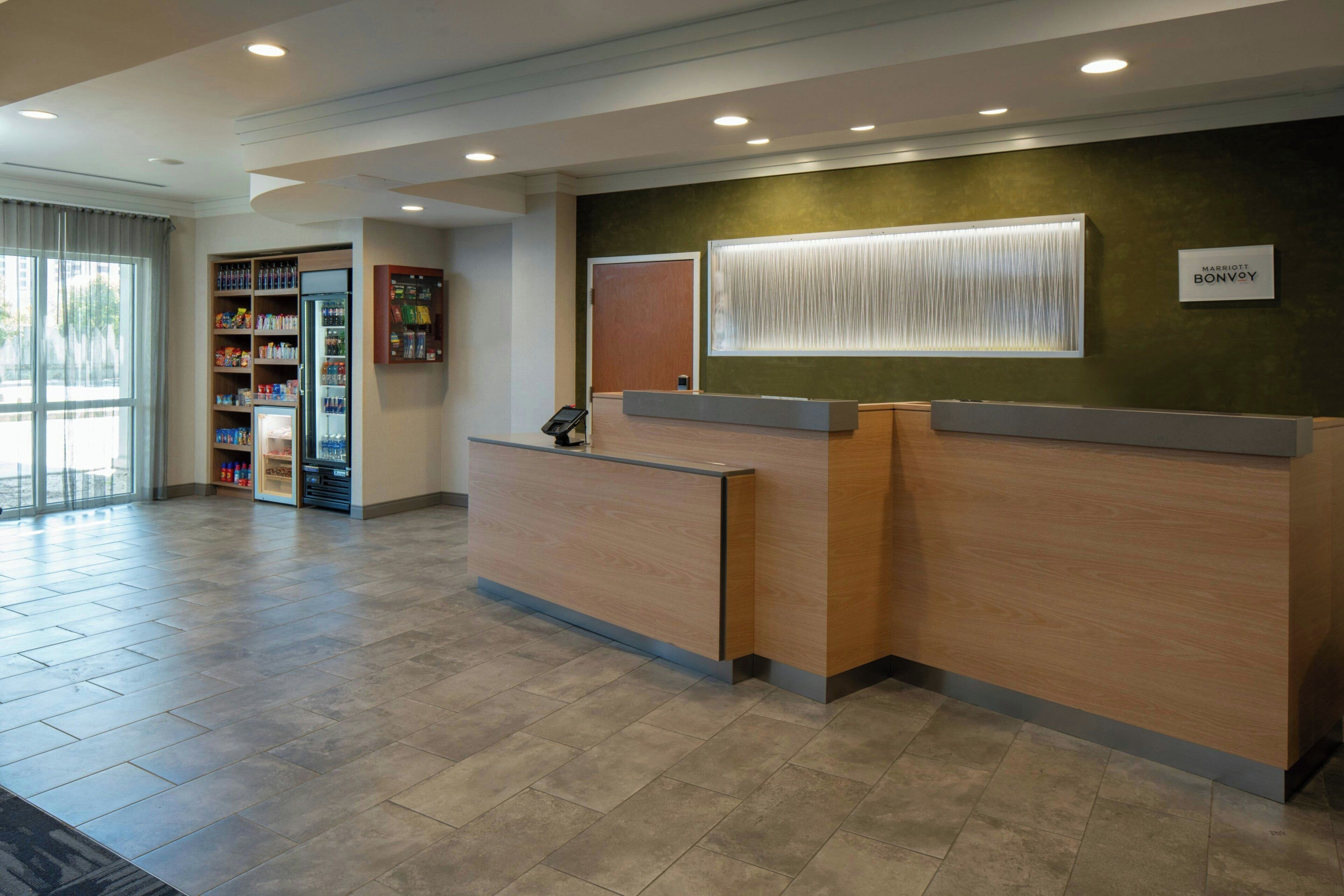 A hotel reception area with a wooden reception desk, a vending machine, and a large window with blinds.