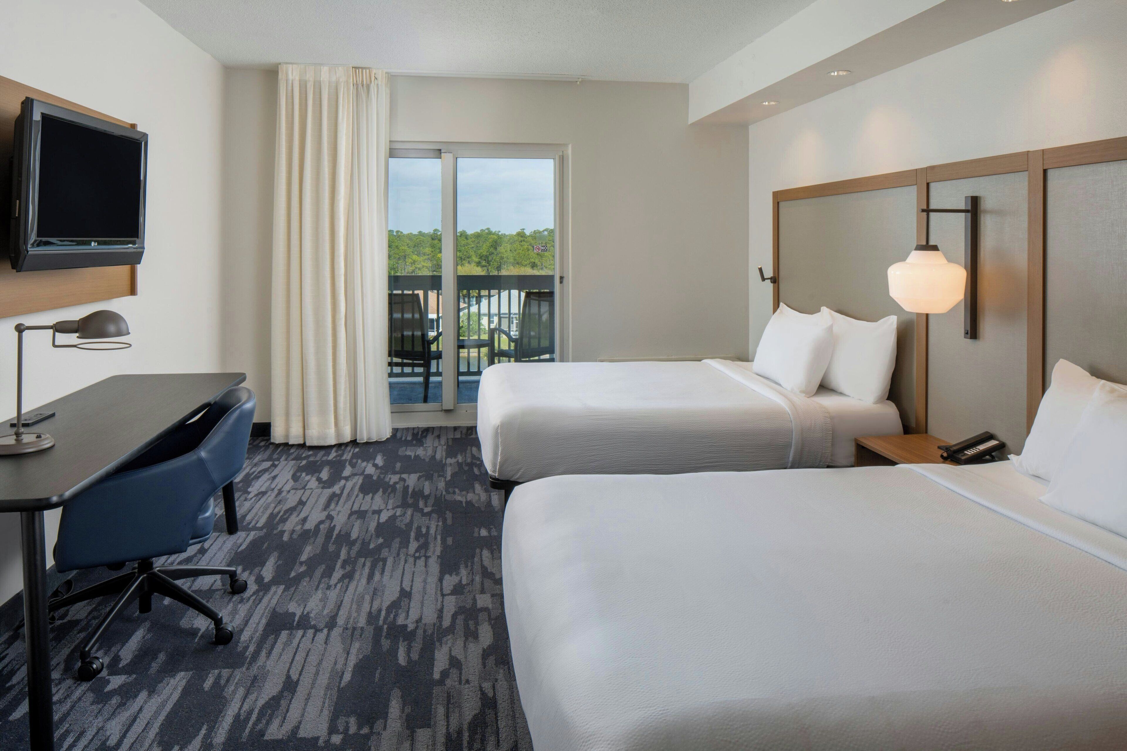 A hotel room with two beds, a desk, a TV, and a balcony with a view.
