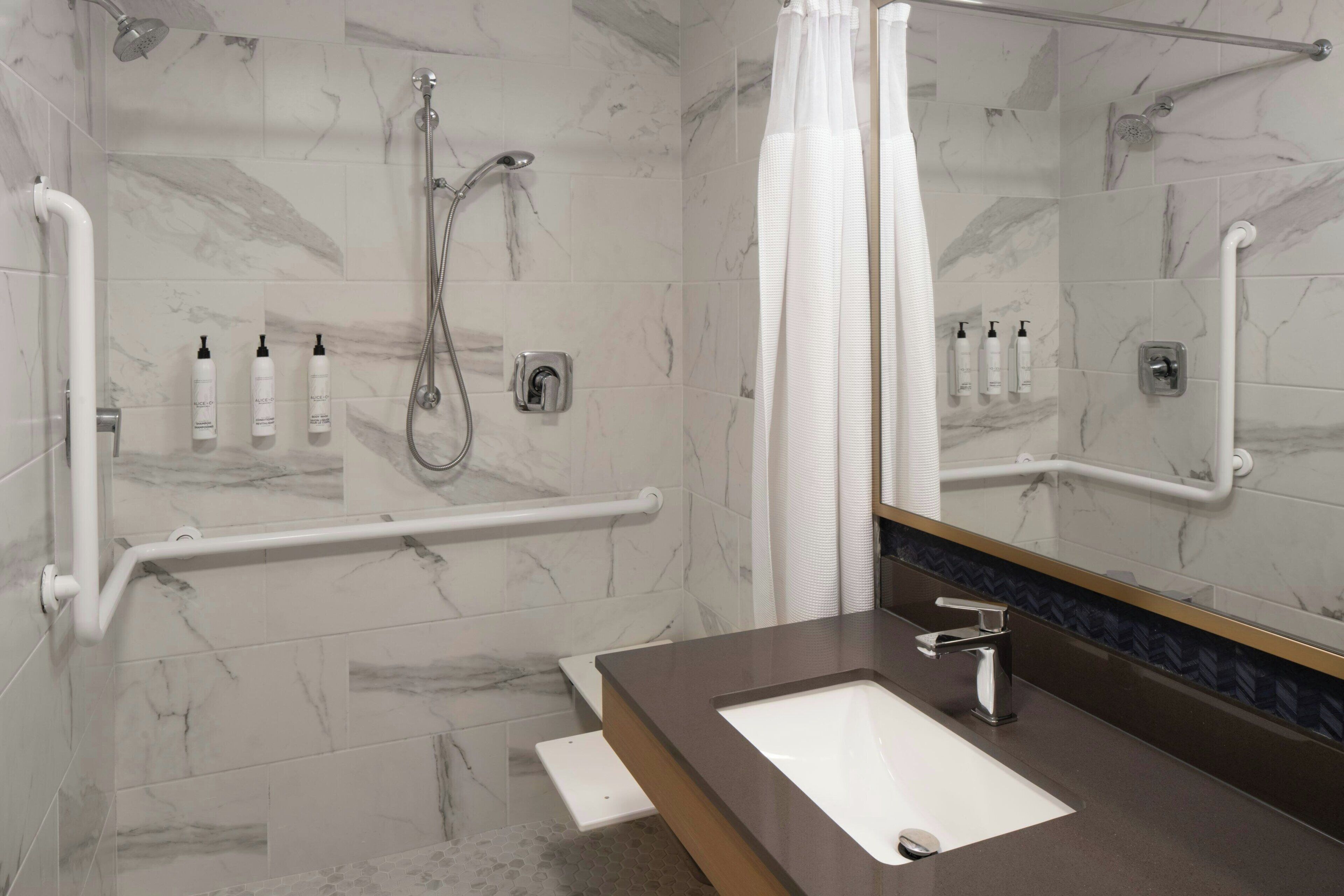 Un baño moderno con piso de mármol, lavamanos blanco, espejo y zona de ducha con barras de apoyo.