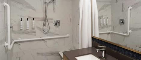 Un baño moderno con piso de mármol, lavamanos blanco, espejo y zona de ducha con barras de apoyo.