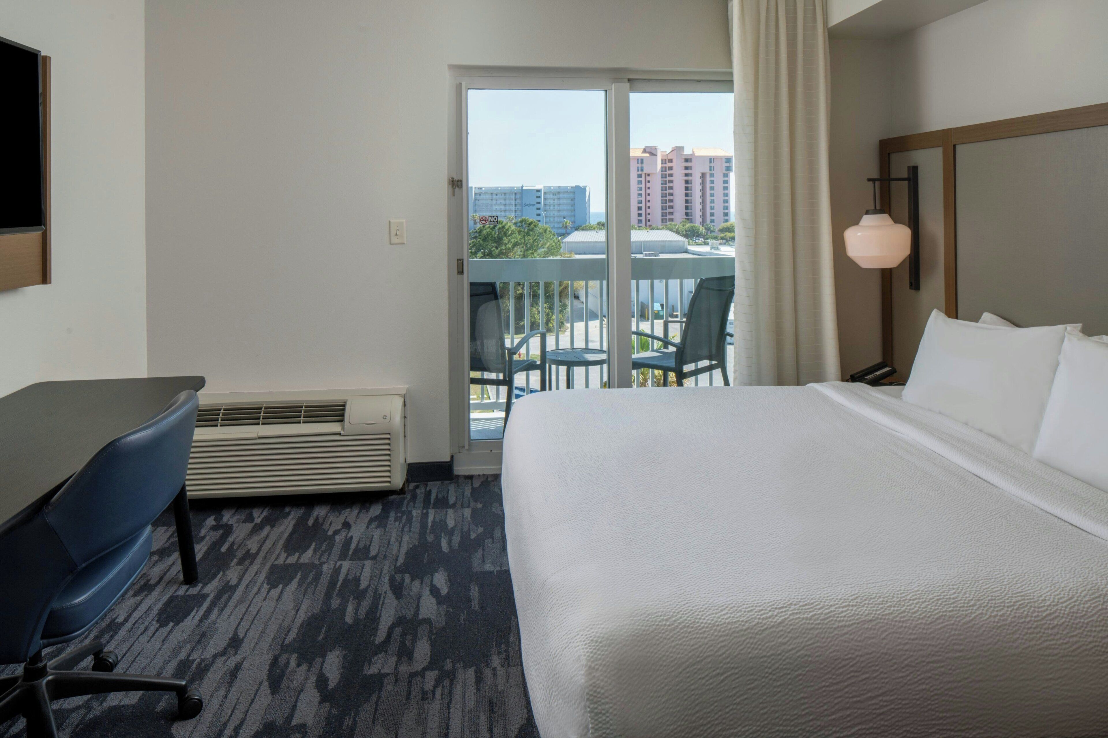 A hotel room with a bed, a desk, a chair, a balcony with a view of buildings, and a hanging light fixture.