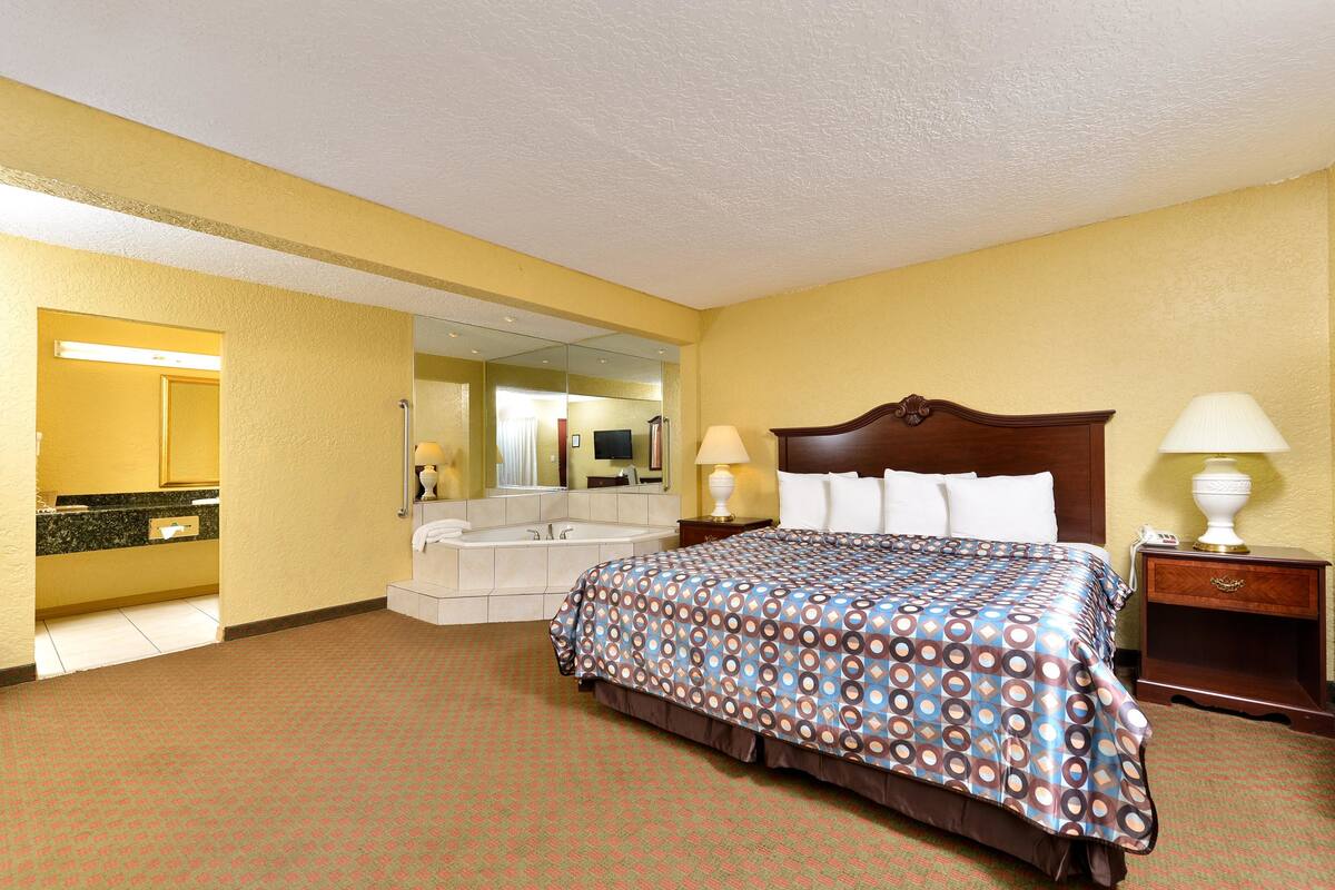 A hotel room with a bed, bedside lamps, a nightstand, a mirror, a bathroom with a sink and a bathtub.